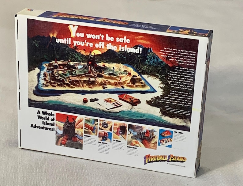 Może przedstawiać: Pudełko gry planszowej Fireball Island. Pudełko zawiera kolorową ilustrację wyspy z tekstem "You won't be safe until you're off the island!" i "A Whole World of Island Adventures!"
