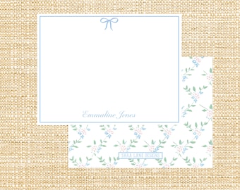 May include: A white card with a blue ribbon at the top and the text "Emmaline Jones" at the bottom. The card is on a light brown burlap background. The card is partially obscured by a white background with a blue and pink floral pattern. The floral pattern has a small text box at the bottom that reads "Sara Lane Designs".