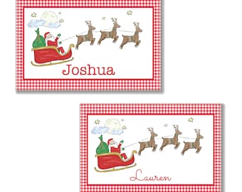 May include: Two rectangular Christmas-themed cards with a red and white gingham border. Each card features Santa Claus in a red sleigh pulled by reindeer under a full moon. The name "Joshua" and "Lauren" are written on the cards.
