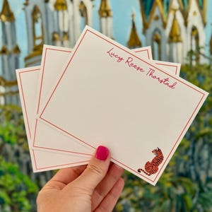 May include: White stationery with a red border and the name "Lucy Reese Thorstad" in pink script. A small orange and black tiger illustration is in the corner. The stationery is held in front of a colorful background.
