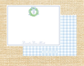 May include: A white card with a blue border and a blue and green wreath with the letter "B" in the center. The text "Emmeline Grace Burke" is written in blue cursive at the bottom of the card. The card is on a brown burlap background. A blue and white gingham pattern is visible in the bottom right corner of the image. The text "SARA LANE DESIGNS" is printed in black on a white rectangle in the bottom right corner of the image.