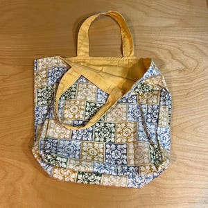 Pode incluir: Uma sacola de tecido com um padr&atilde;o repetitivo de azulejos quadrados em tons de azul, verde e bege. A bolsa possui duas al&ccedil;as de tecido amarelo e um forro interno amarelo correspondente, feita de um material t&ecirc;xtil macio e vis&iacute;vel.