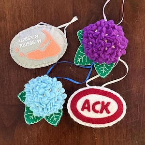 May include: A collection of felt ornaments. One is a gray oval with a peach-colored map and coordinates. Another is a purple hydrangea with green leaves. A blue hydrangea and a red and white oval with "ACK" are also shown.