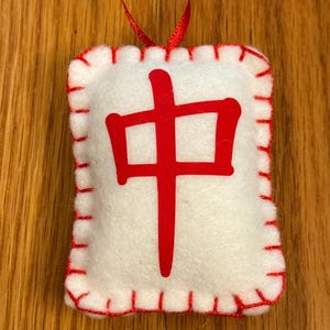 May include: A white felt ornament with a red stitched border and a red Chinese character in the center. A red ribbon is attached for hanging. The ornament is rectangular in shape.
