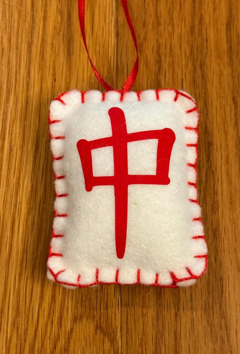 May include: A white felt ornament with a red stitched border and a red Chinese character in the center. A red ribbon is attached for hanging. The ornament is rectangular in shape.