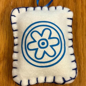 May include: A white felt ornament with a blue floral design inside a circle. The edges are stitched with blue thread. A blue ribbon is attached for hanging. The ornament is rectangular in shape.
