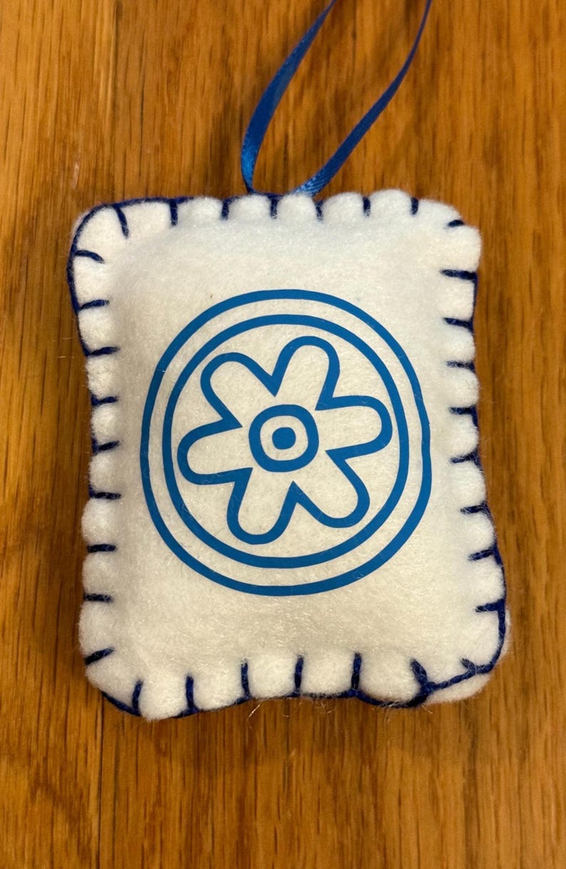 May include: A white felt ornament with a blue floral design inside a circle. The edges are stitched with blue thread. A blue ribbon is attached for hanging. The ornament is rectangular in shape.