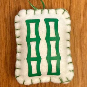 May include: A white felt ornament with a green bamboo design. The ornament is rectangular with rounded corners and green stitching around the edges. A green ribbon is attached for hanging.