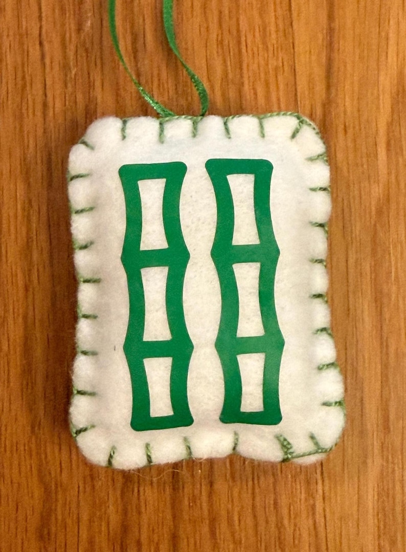 May include: A white felt ornament with a green bamboo design. The ornament is rectangular with rounded corners and green stitching around the edges. A green ribbon is attached for hanging.