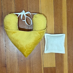 Pode incluir: Uma almofada em forma de cora&ccedil;&atilde;o amarelo-mostarda com um padr&atilde;o de favo de mel em relevo e um motivo de abelha, acentuada por fitas brancas no topo. Ao lado, encontra-se uma pequena bolsa quadrada de tecido branco liso com bordas costuradas.