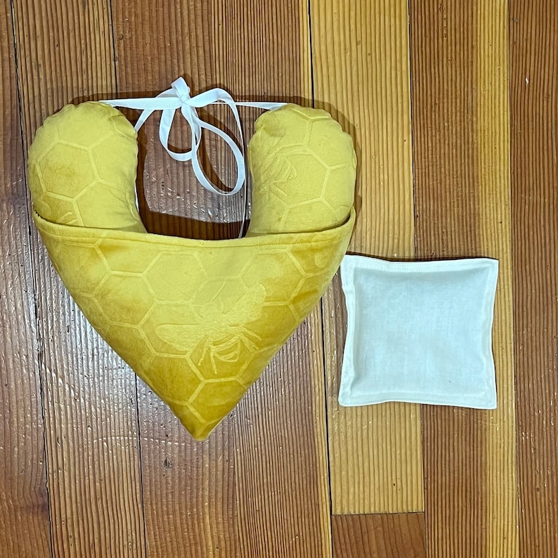 Pode incluir: Uma almofada em forma de cora&ccedil;&atilde;o amarelo-mostarda com um padr&atilde;o de favo de mel em relevo e um motivo de abelha, acentuada por fitas brancas no topo. Ao lado, encontra-se uma pequena bolsa quadrada de tecido branco liso com bordas costuradas.
