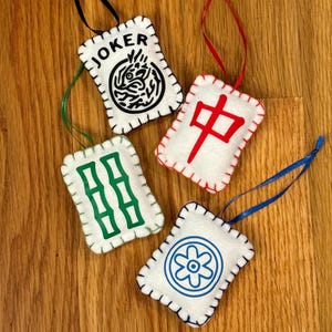May include: Four white felt ornaments with stitched edges and hanging ribbons. One ornament has the word "JOKER" and a black design. Another has a red Chinese character. The other two have green and blue designs. These are likely Mahjong game pieces.