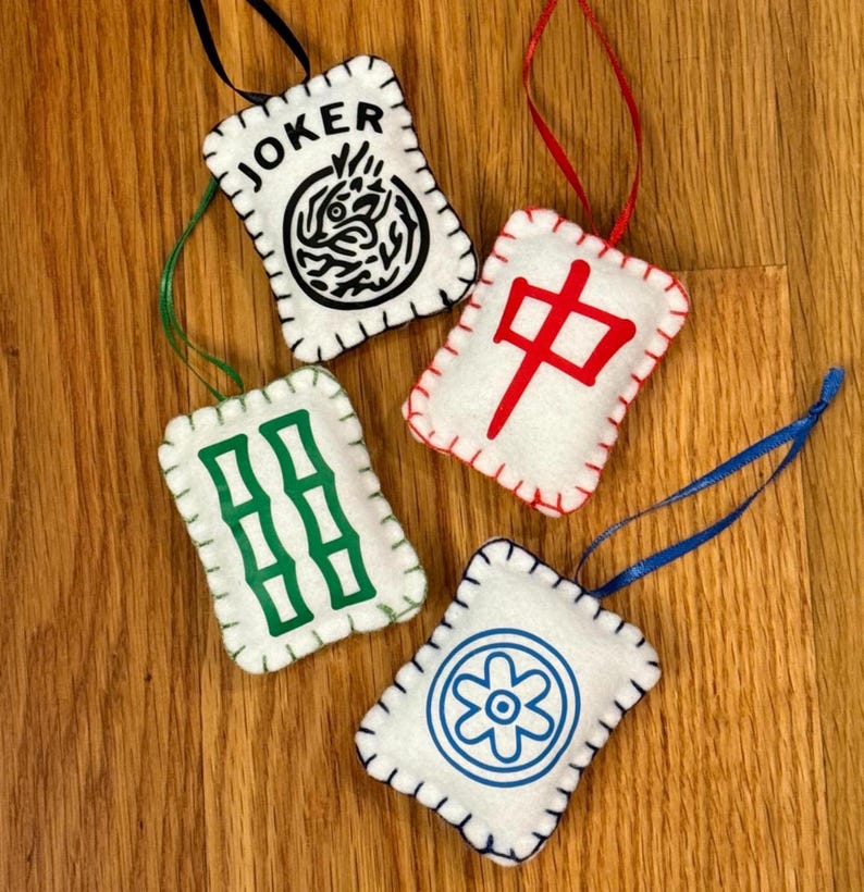 May include: Four white felt ornaments with stitched edges and hanging ribbons. One ornament has the word "JOKER" and a black design. Another has a red Chinese character. The other two have green and blue designs. These are likely Mahjong game pieces.
