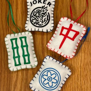 May include: Four white felt ornaments with stitched edges and ribbon hangers. Each ornament features a different design: "JOKER" with a black graphic, green bamboo, a red Chinese character, and a blue floral design. The ornaments are on a wooden surface.