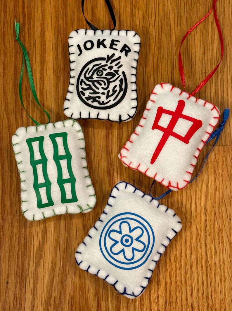 May include: Four white felt ornaments with stitched edges and ribbon hangers. Each ornament features a different design: "JOKER" with a black graphic, green bamboo, a red Chinese character, and a blue floral design. The ornaments are on a wooden surface.