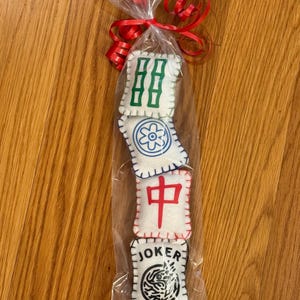 May include: A clear cellophane bag tied with a red ribbon, containing four white felt Mahjong tiles. The tiles have green, blue, and red symbols, and one tile reads "JOKER" with a black and white design.