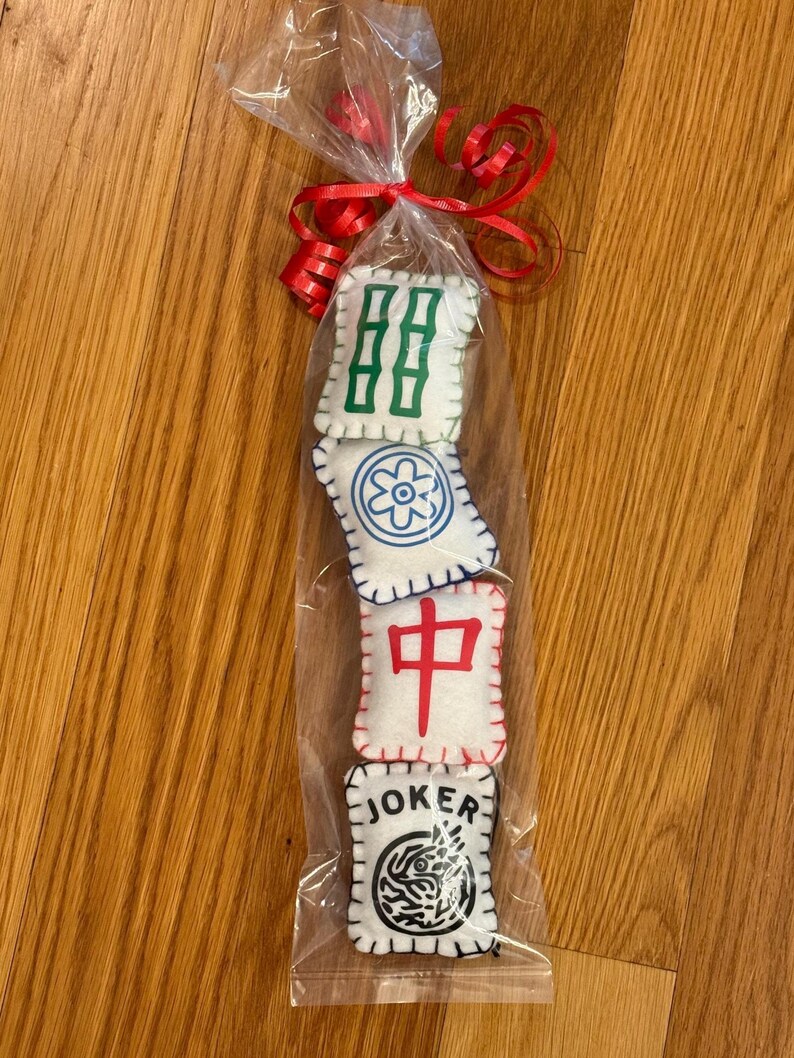 May include: A clear cellophane bag tied with a red ribbon, containing four white felt Mahjong tiles. The tiles have green, blue, and red symbols, and one tile reads "JOKER" with a black and white design.