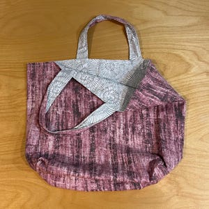 Pode incluir: Uma bolsa tote revers&iacute;vel com uma estampa abstrata texturizada em malva escuro e preto de um lado e um padr&atilde;o circular de raios de sol em cinza claro e branco do outro. A bolsa inclui duas al&ccedil;as de tecido e um corpo retangular aberto.