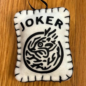 May include: A white felt ornament with the word "JOKER" in black block letters. Below the text is a black circular design. The ornament has a black stitched border and a black ribbon for hanging.