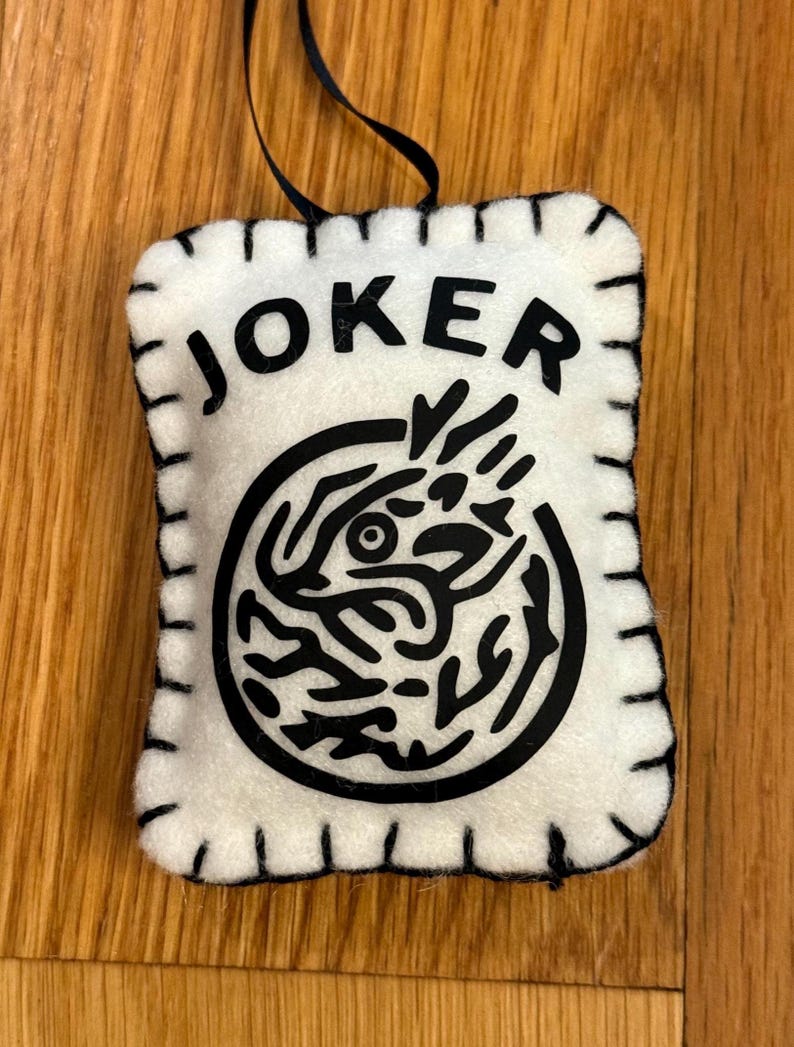 May include: A white felt ornament with the word "JOKER" in black block letters. Below the text is a black circular design. The ornament has a black stitched border and a black ribbon for hanging.