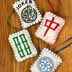 May include: Four white felt ornaments with stitched edges and ribbon hangers. Each ornament features a different design in black, green, red, and blue. One says "JOKER" with a black graphic. Others have Chinese symbols and a flower.