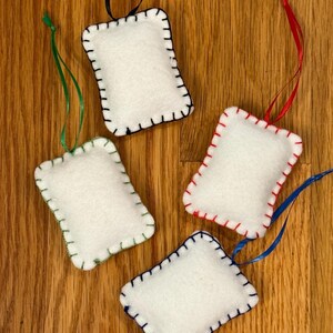 May include: Four white felt ornaments with colorful stitching and ribbon hangers. Each ornament is rectangular with rounded corners and a different color of stitching: black, green, red, and blue. The ribbons are black, green, red, and blue.