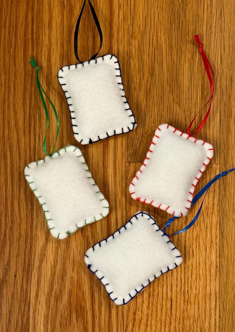 May include: Four white felt ornaments with colorful stitching and ribbon hangers. Each ornament is rectangular with rounded corners and a different color of stitching: black, green, red, and blue. The ribbons are black, green, red, and blue.