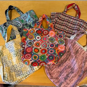 Pode incluir: Cinco sacos de tecido com v&aacute;rios padr&otilde;es, incluindo uma estampa paisley verde e azul, um xadrez vermelho e preto, um design de azulejos amarelo e branco, uma estampa floral circular multicolorida e um tecido rosa empoeirado com duas al&ccedil;as.