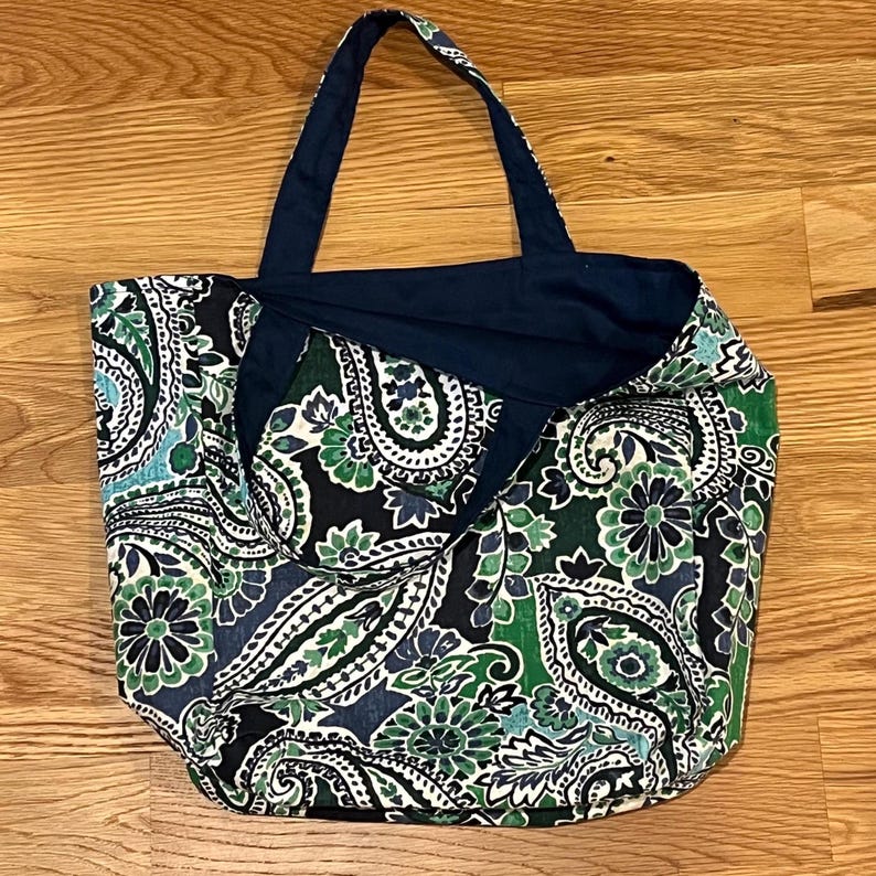 Pode incluir: Uma bolsa tote com um padr&atilde;o paisley em azul escuro, verde e branco. A bolsa tem duas al&ccedil;as de tecido azul escuro e um forro interior azul escuro s&oacute;lido. O tecido exterior apresenta um design denso e repetitivo de flores e paisley.