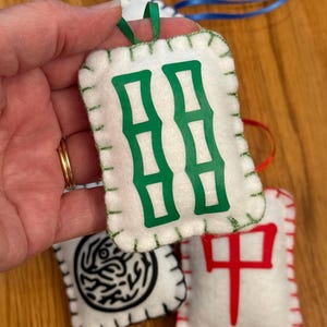 May include: Handmade felt Mahjong tile ornaments. The white felt tiles feature green, black, and red symbols, with green and red stitching around the edges. Each tile has a ribbon loop for hanging.