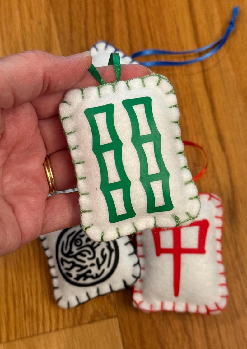 May include: Handmade felt Mahjong tile ornaments. The white felt tiles feature green, black, and red symbols, with green and red stitching around the edges. Each tile has a ribbon loop for hanging.