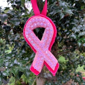 Pode incluir: Um ornamento de fita de consciencializa&ccedil;&atilde;o rosa feito de feltro, com um design de camada dupla com um verso rosa brilhante e uma frente rosa mais clara. A frente &eacute; adornada com uma &uacute;nica fila de contas transparentes, facetadas e iridescentes.