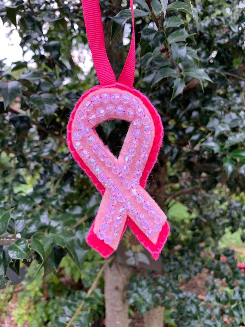 Pode incluir: Um ornamento de fita de consciencializa&ccedil;&atilde;o rosa feito de feltro, com um design de camada dupla com um verso rosa brilhante e uma frente rosa mais clara. A frente &eacute; adornada com uma &uacute;nica fila de contas transparentes, facetadas e iridescentes.
