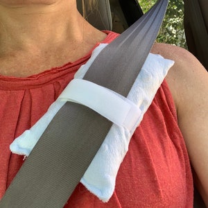 Pode incluir: Uma capa de cinto de seguran&ccedil;a azul-clara com textura macia e uma tira de fecho de velcro branca. A almofada retangular est&aacute; enrolada em torno de um cinto de seguran&ccedil;a cinzento, proporcionando uma camada acolchoada sobre o peito.