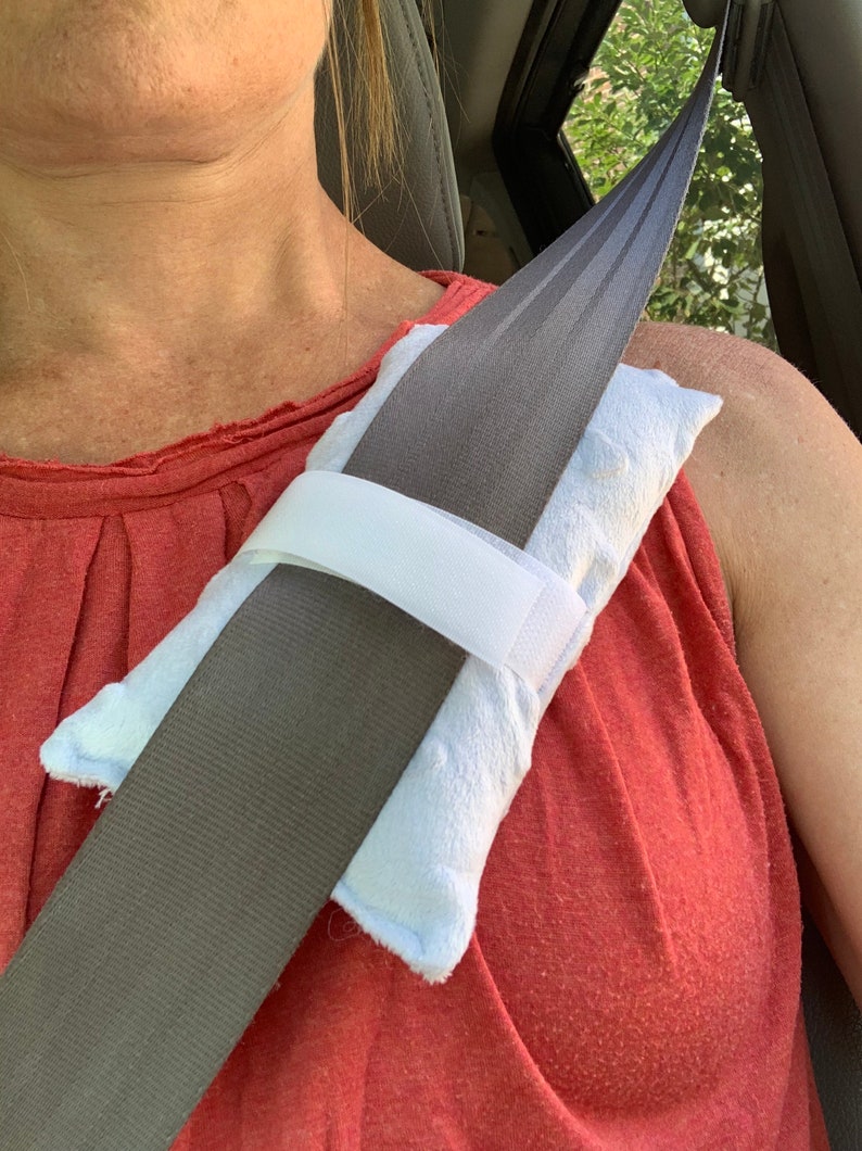 Pode incluir: Uma capa de cinto de seguran&ccedil;a azul-clara com textura macia e uma tira de fecho de velcro branca. A almofada retangular est&aacute; enrolada em torno de um cinto de seguran&ccedil;a cinzento, proporcionando uma camada acolchoada sobre o peito.