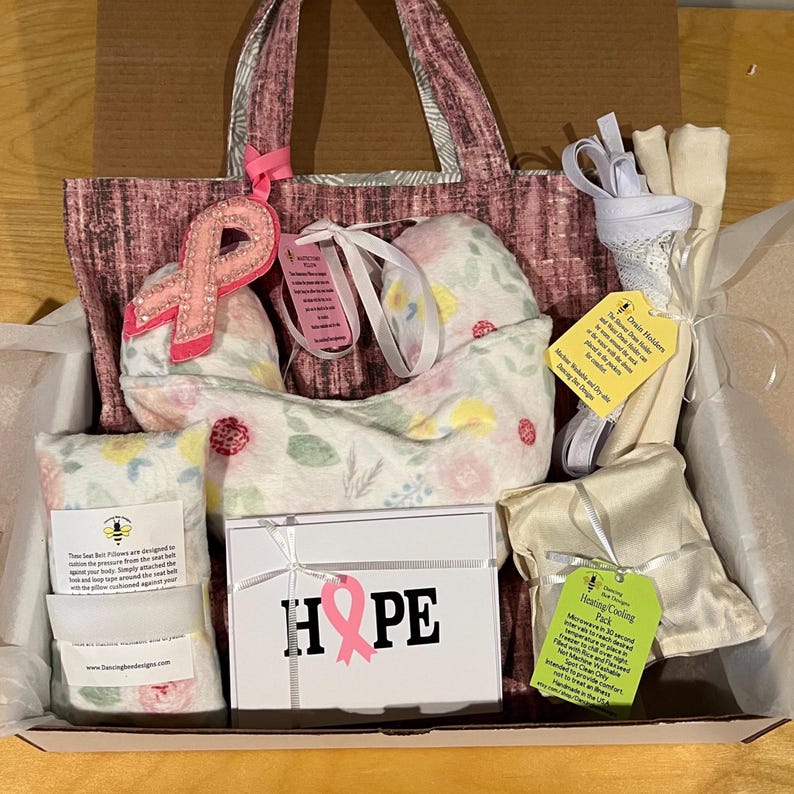 Pode incluir: Um pacote de cuidados contendo uma bolsa rosa estampada, uma almofada de pesco&ccedil;o floral com um broche de fita rosa, uma caixa branca com o r&oacute;tulo HOPE e uma fita rosa, uma bolsa t&eacute;rmica e suportes de drenagem brancos com etiquetas informativas.