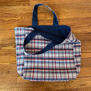 Pode incluir: Uma bolsa tote revers&iacute;vel com um padr&atilde;o xadrez de um lado, com linhas vermelhas, brancas, azuis e pretas. O lado oposto da bolsa &eacute; de tecido azul-marinho s&oacute;lido. A bolsa inclui duas al&ccedil;as de ombro a condizer em tecido azul-marinho.
