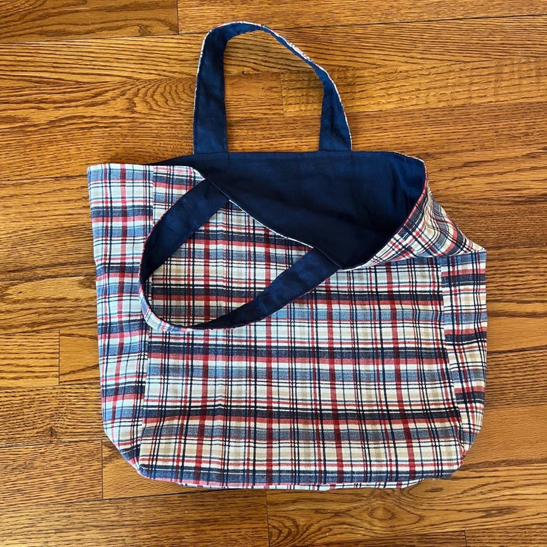 Pode incluir: Uma bolsa tote revers&iacute;vel com um padr&atilde;o xadrez de um lado, com linhas vermelhas, brancas, azuis e pretas. O lado oposto da bolsa &eacute; de tecido azul-marinho s&oacute;lido. A bolsa inclui duas al&ccedil;as de ombro a condizer em tecido azul-marinho.