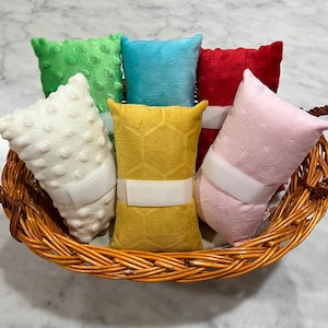 Pode incluir: Uma cole&ccedil;&atilde;o de seis almofadas retangulares de pel&uacute;cia nas cores branca, verde, azul, vermelha, amarela e rosa, dispostas numa cesta de vime tran&ccedil;ado castanho. As almofadas apresentam texturas variadas, incluindo pontos em relevo e padr&otilde;es.