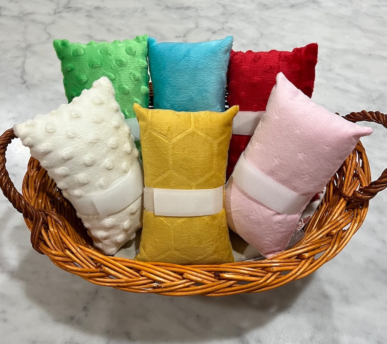 Pode incluir: Uma cole&ccedil;&atilde;o de seis almofadas retangulares de pel&uacute;cia nas cores branca, verde, azul, vermelha, amarela e rosa, dispostas numa cesta de vime tran&ccedil;ado castanho. As almofadas apresentam texturas variadas, incluindo pontos em relevo e padr&otilde;es.
