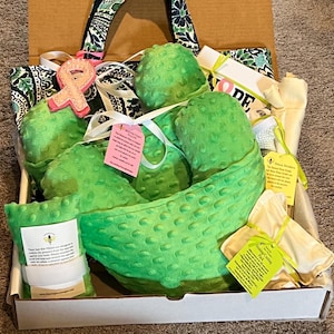 Pode incluir: Um pacote de cuidados contendo uma almofada de mastectomia de tecido minky verde brilhante texturizado, uma almofada de cinto de seguran&ccedil;a, uma bolsa t&eacute;rmica e um suporte de drenagem, com etiquetas informativas e um broche de fita rosa.