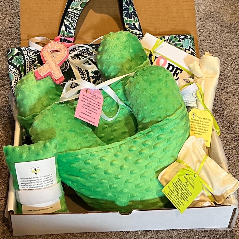 Pode incluir: Um pacote de cuidados contendo uma almofada de mastectomia de tecido minky verde brilhante texturizado, uma almofada de cinto de seguran&ccedil;a, uma bolsa t&eacute;rmica e um suporte de drenagem, com etiquetas informativas e um broche de fita rosa.