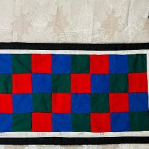 May include: A colorful, quilted table runner with a checkerboard pattern of red, blue, and green squares. The runner has a white border, a black outer frame, and two black fabric pieces attached to the side.