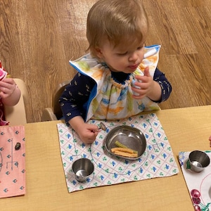 May include: A child wearing a bib with a blue trim and a floral pattern sits at a table with a placemat and a small metal bowl. The bowl contains food. Another child is visible at the table, also wearing a bib and eating.