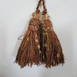 May include: Two brown and gold tassel tiebacks with clear glass beads. The tassels are made of a woven fabric with a fringe.