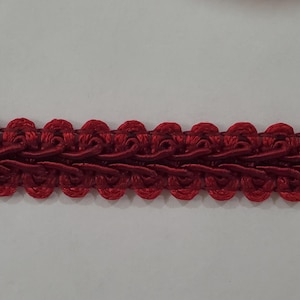May include: A length of decorative red trim with a repeating floral design. The trim is made of a woven material and features a scalloped edge. The color is a deep, rich red, and the trim appears to be new.