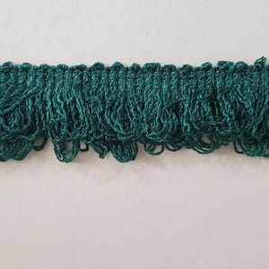 May include: A length of dark teal fringe trim. The trim has a woven top edge and a series of looped, knotted strands that hang down. The fringe is a solid color and appears to be made of a soft, textured material.