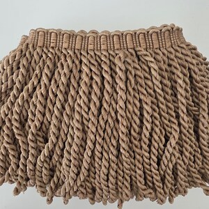 May include: Close-up of a beige rope lampshade with a fringed bottom. The lampshade has a woven top section and long, twisted rope fringe. The neutral color and texture suggest a bohemian or coastal decor style.