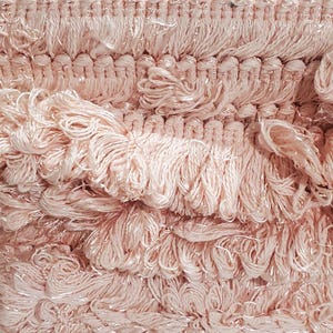 May include: Close-up of a roll of pale pink fringe trim. The trim features a textured design with looped and fringed elements. The material appears to be a soft, possibly synthetic fabric, suitable for various craft and sewing projects.