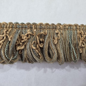May include: Decorative fringe trim in shades of tan, gray, and light blue. The trim features a woven top edge with looped and fringed elements, adding texture and visual interest. Ideal for upholstery and craft projects.